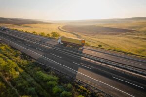 A large truck drives down a multi-lane highway through open countryside at sunset, surrounded by green fields and gentle hills under a bright sky.