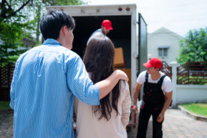 A couple stands arm in arm, watching as movers from Scarborough Moving Services in red caps load boxes into a moving truck parked in a driveway on a sunny day.