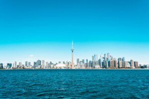 Toronto skyline with the CN Tower and surrounding skyscrapers seen across a large body of blue water under a clear, bright blue sky.