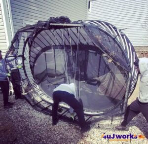 Four people are attempting to move or reposition a large, circular trampoline that is bent and damaged, situated next to a house. The trampolines frame appears twisted and misshapen.
