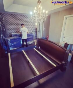 A person in a 4Uworks Moving Company shirt wraps furniture in plastic in a room with a chandelier and a partially assembled dark wood bed frame.