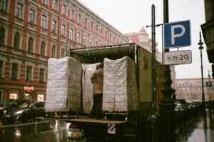 A person stands in the back of a moving service in North Yorks delivery truck, arranging large, padded items on a rainy city street as cars pass by and a blue parking sign is visible nearby.
