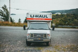 A white U-Haul moving truck is parked on a gravel lot with trees and hills in the background under a cloudy sky.
