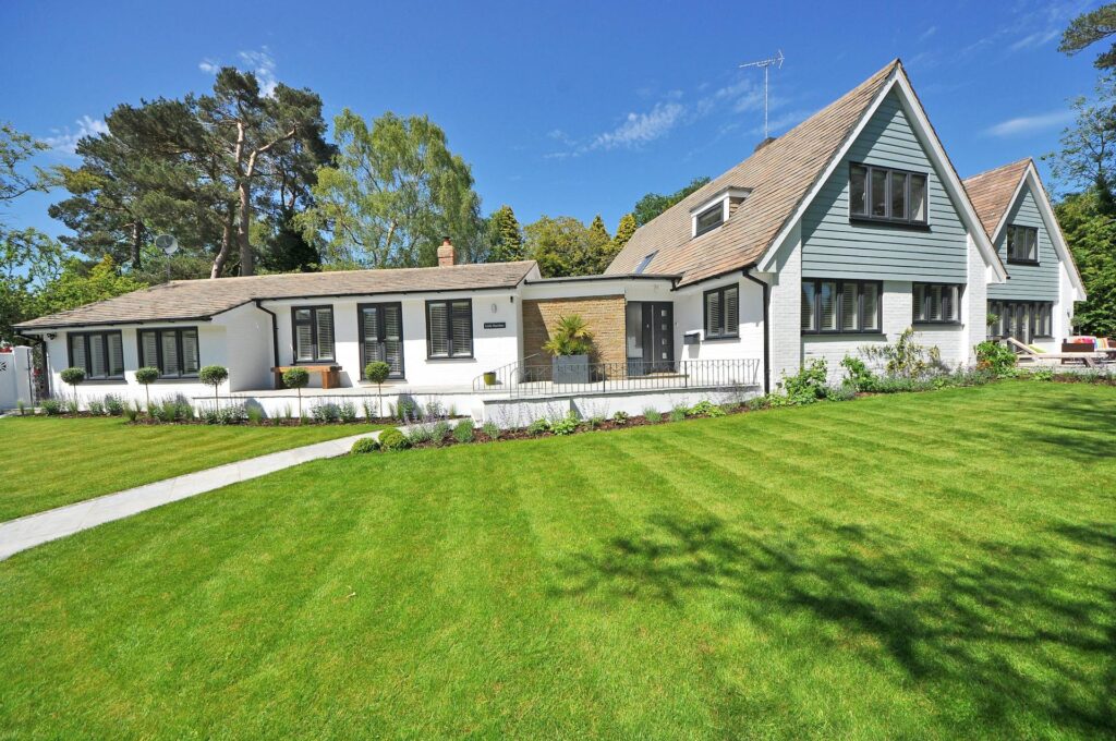 A modern house with white walls, large windows, and a well-manicured green lawn under a clear blue sky. Trees and shrubs surround the property, creating a peaceful suburban setting.