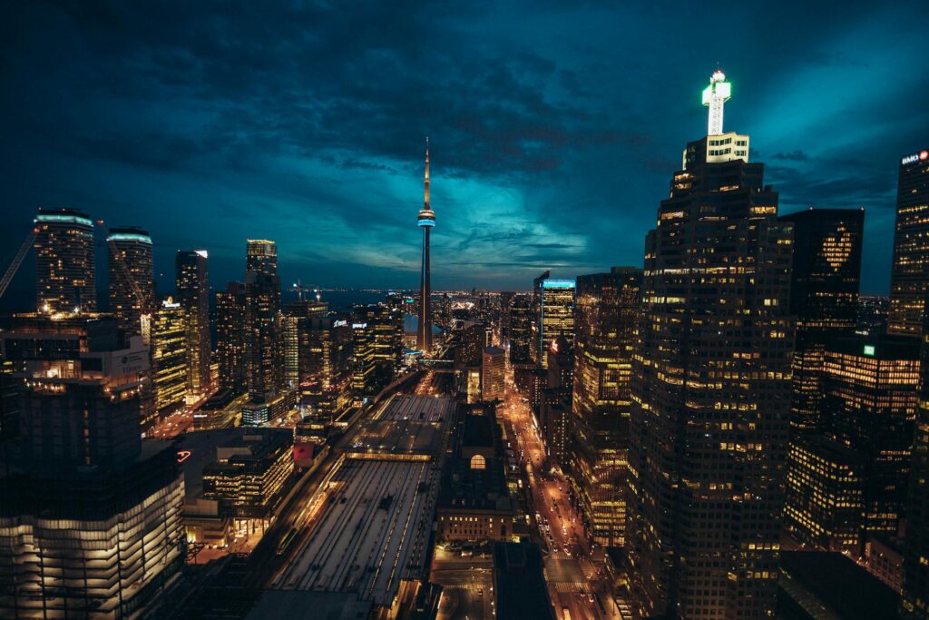A nighttime cityscape of downtown Toronto, featuring the brightly lit CN Tower and illuminated skyscrapers under a dark blue, cloudy sky. Streets and buildings glow with yellow and white lights.